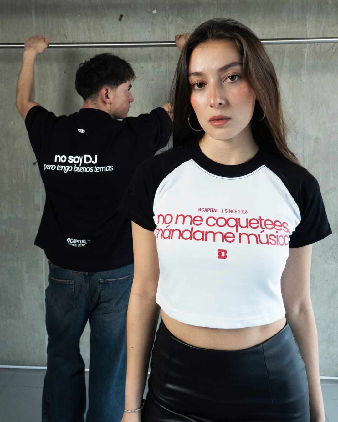 Ella de frente con la baby tee blanca de mangas negras y la frase «no me coquetees, mándame música»; detrás, él de espaldas con la oversize negra «no soy DJ pero tengo buenos temas».
